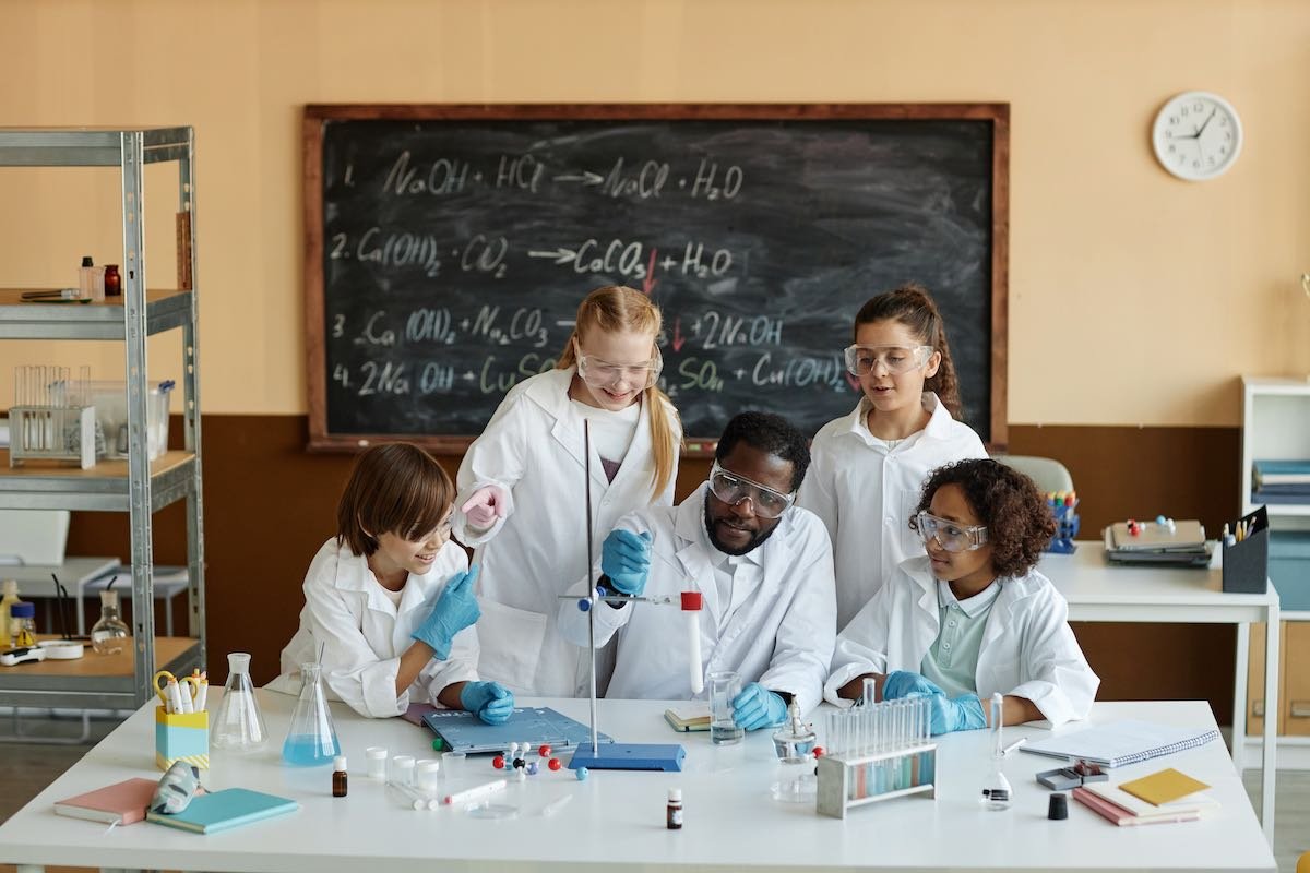 A science teacher in a lab coat works on a science experiment at a desk with a group of diverse young teens.